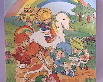 Puede incluir: Caja de rompecabezas vintage de 200 piezas con una colorida ilustración de un arcoíris, un unicornio blanco y personajes de dibujos animados. Los personajes visten varios atuendos en un paisaje caprichoso.
