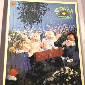 May include: A 25-piece Cabbage Patch Kids puzzle featuring four dolls in a red wagon. The puzzle has a blue sky background with a tree and flowers. The puzzle is for ages 3 to 7.