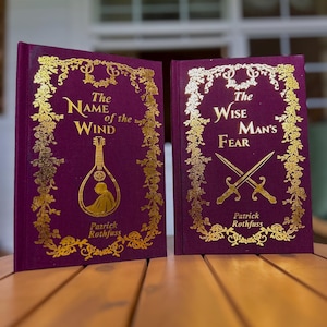 Pode incluir: Dois livros de capa dura com capas bord&ocirc; e letras douradas. O primeiro livro &eacute; intitulado "The Name of the Wind" e apresenta uma ilustra&ccedil;&atilde;o de ala&uacute;de. O segundo livro &eacute; intitulado "The Wise Man's Fear" com espadas cruzadas.