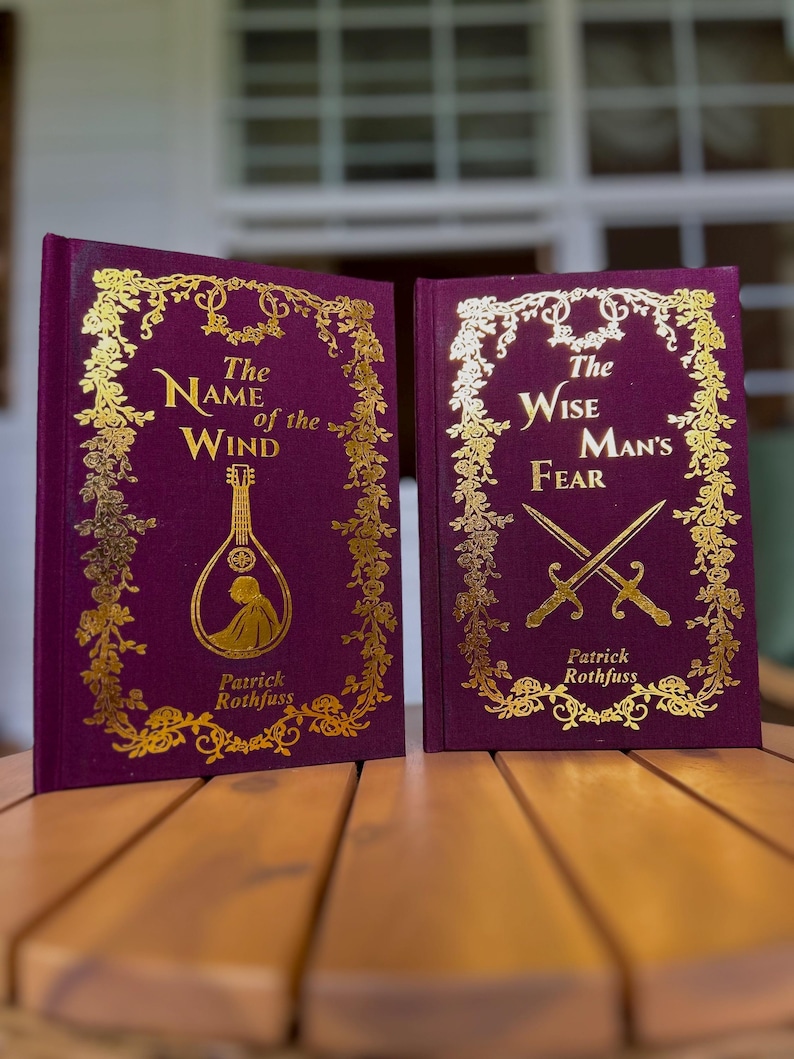 Pode incluir: Dois livros de capa dura com capas bord&ocirc; e letras douradas. O primeiro livro &eacute; intitulado "The Name of the Wind" e apresenta uma ilustra&ccedil;&atilde;o de ala&uacute;de. O segundo livro &eacute; intitulado "The Wise Man's Fear" com espadas cruzadas.