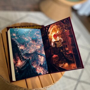 Pode incluir: Um livro aberto com ilustra&ccedil;&otilde;es de fantasia. Uma p&aacute;gina mostra uma cidade com luzes, a outra uma pessoa lendo em uma biblioteca. O livro tem uma capa bord&ocirc; e est&aacute; em uma mesa de madeira.