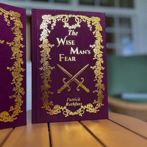 Pode incluir: Um livro de capa dura intitulado "The Wise Man's Fear" de Patrick Rothfuss. O livro tem uma capa roxa escura com letras douradas e detalhes decorativos florais e de espadas. O livro &eacute; exibido em uma superf&iacute;cie de madeira.