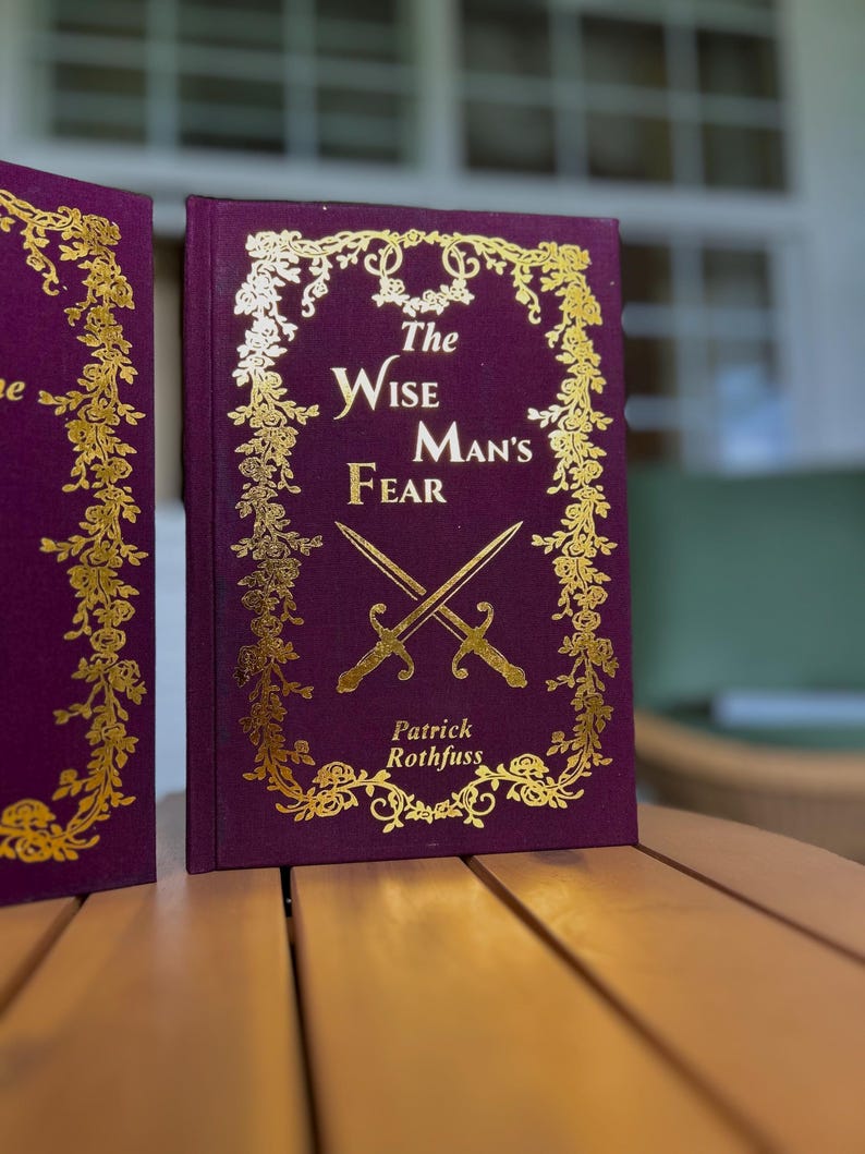 Pode incluir: Um livro de capa dura intitulado "The Wise Man's Fear" de Patrick Rothfuss. O livro tem uma capa roxa escura com letras douradas e detalhes decorativos florais e de espadas. O livro &eacute; exibido em uma superf&iacute;cie de madeira.