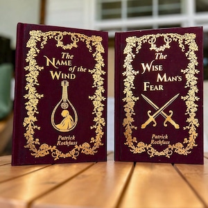 Pode incluir: Dois livros de capa dura com capas bord&ocirc; e letras e desenhos dourados. O primeiro livro &eacute; intitulado "The Name of the Wind" e o segundo "The Wise Man's Fear". Ambos os livros s&atilde;o de Patrick Rothfuss.