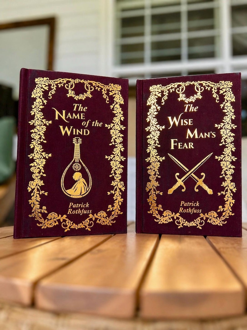 Pode incluir: Dois livros de capa dura com capas bord&ocirc; e letras e desenhos dourados. O primeiro livro &eacute; intitulado "The Name of the Wind" e o segundo "The Wise Man's Fear". Ambos os livros s&atilde;o de Patrick Rothfuss.