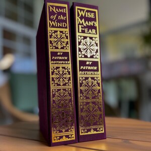 Pode incluir: Dois livros de capa dura com capas bord&ocirc; e letras e desenhos dourados em relevo. As lombadas dizem "Name of the Wind" e "Wise Man's Fear" de Patrick Rothfuss. Exibidos em uma superf&iacute;cie de madeira.