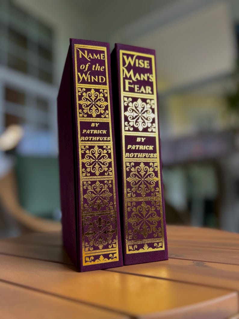 Pode incluir: Dois livros de capa dura com capas bord&ocirc; e letras e desenhos dourados em relevo. As lombadas dizem "Name of the Wind" e "Wise Man's Fear" de Patrick Rothfuss. Exibidos em uma superf&iacute;cie de madeira.