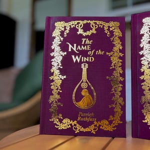 Pode incluir: Um livro de capa dura intitulado "The Name of the Wind" de Patrick Rothfuss. O livro tem uma capa roxa escura com letras douradas e detalhes decorativos. Uma ilustra&ccedil;&atilde;o dourada de um ala&uacute;de &eacute; apresentada na capa.