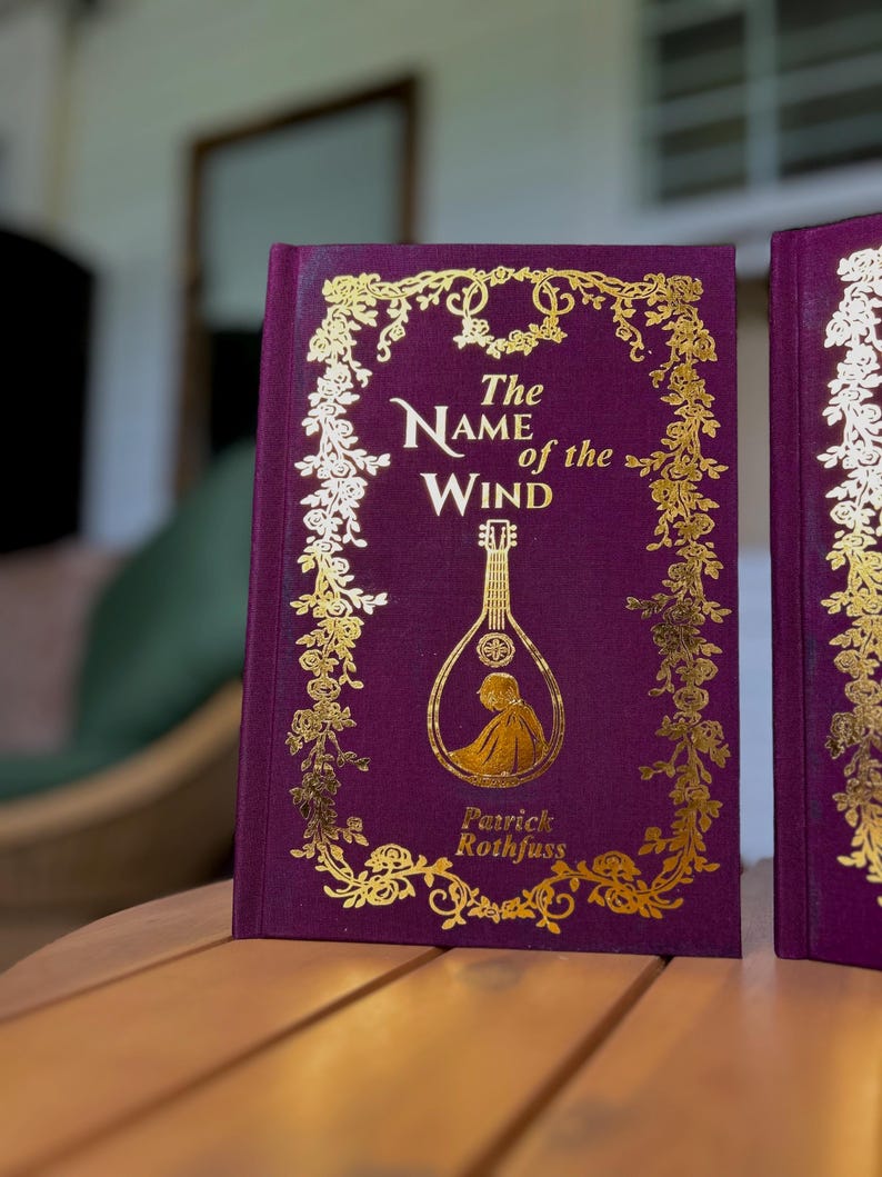 Pode incluir: Um livro de capa dura intitulado "The Name of the Wind" de Patrick Rothfuss. O livro tem uma capa roxa escura com letras douradas e detalhes decorativos. Uma ilustra&ccedil;&atilde;o dourada de um ala&uacute;de &eacute; apresentada na capa.