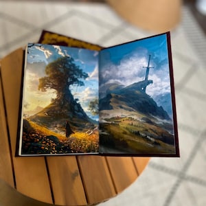 Pode incluir: Um livro aberto com duas ilustra&ccedil;&otilde;es de paisagens. Uma mostra uma grande &aacute;rvore numa colina, a outra uma montanha com uma espada. O livro tem uma capa bord&ocirc; e est&aacute; numa mesa de madeira.