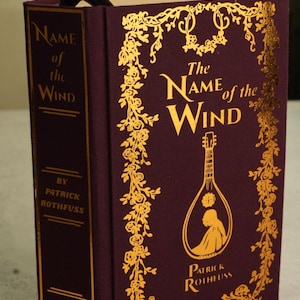 Pode incluir: Um livro de capa dura intitulado "The Name of the Wind" de Patrick Rothfuss. A capa bord&ocirc; apresenta letras douradas e enfeites florais. Uma fita preta como marcador de p&aacute;gina &eacute; vis&iacute;vel.