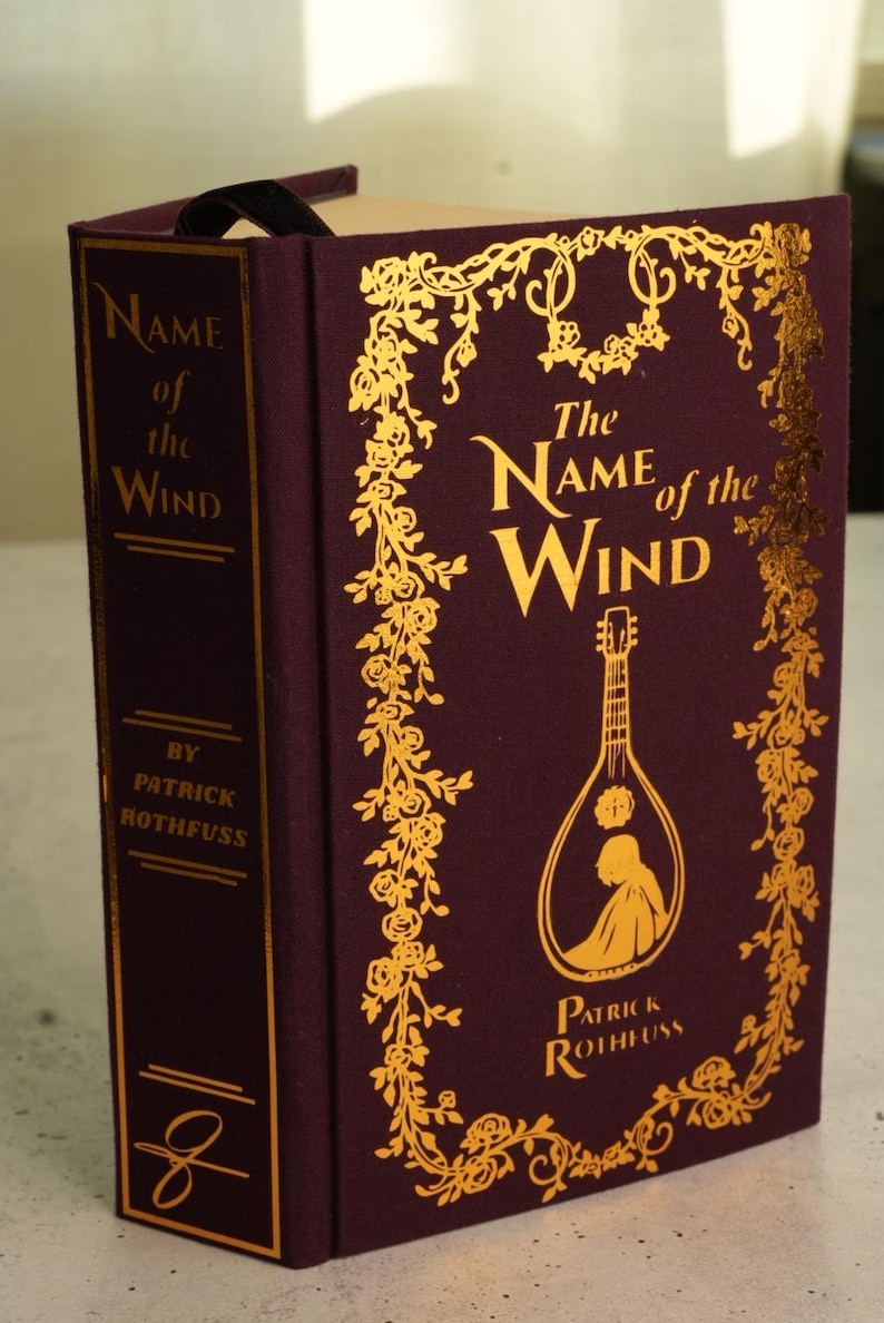 Pode incluir: Um livro de capa dura intitulado "The Name of the Wind" de Patrick Rothfuss. A capa bord&ocirc; apresenta letras douradas e enfeites florais. Uma fita preta como marcador de p&aacute;gina &eacute; vis&iacute;vel.