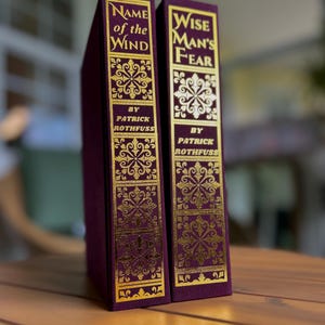 Pode incluir: Dois livros de capa dura com capas bord&ocirc; e letras e desenhos dourados em relevo. As lombadas dizem "Name of the Wind" e "Wise Man's Fear" de Patrick Rothfuss. Os livros s&atilde;o exibidos em uma superf&iacute;cie de madeira.