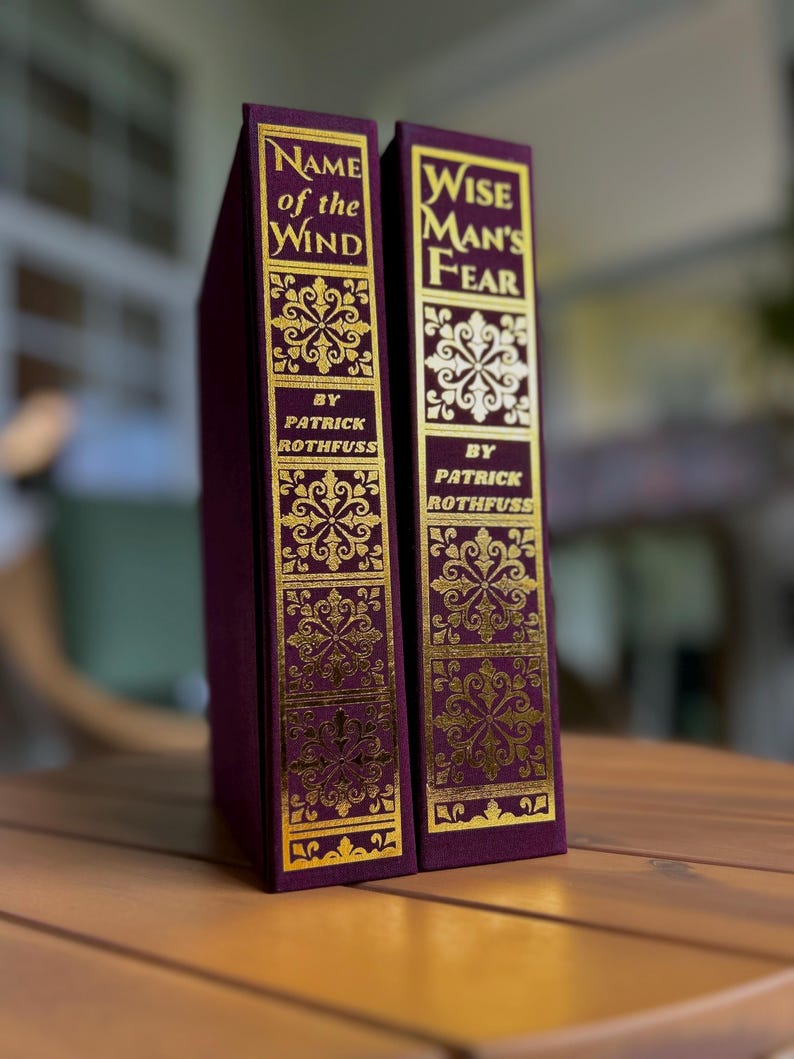 Pode incluir: Dois livros de capa dura com capas bord&ocirc; e letras e desenhos dourados em relevo. As lombadas dizem "Name of the Wind" e "Wise Man's Fear" de Patrick Rothfuss. Os livros s&atilde;o exibidos em uma superf&iacute;cie de madeira.