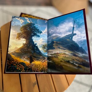 Pode incluir: Um livro aberto com duas ilustra&ccedil;&otilde;es de paisagens. Uma mostra uma pessoa em frente a uma grande &aacute;rvore numa colina, a outra uma espada numa montanha. O livro tem uma capa bord&ocirc; e est&aacute; numa mesa de madeira.
