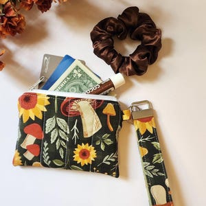 May include: A rectangular fabric zipper pouch with a dark green background featuring a pattern of sunflowers, mushrooms, and leaves. The pouch includes a matching wristlet strap, a white zipper, and contains a credit card, cash, and a lip balm.