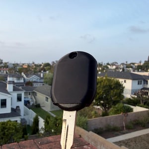 May include: A black car key with a silver metal key blade. The key has a rounded rectangular head with a small hole at the top. The key is set against a background of houses and a blue sky.
