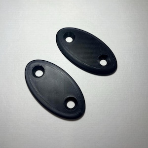 May include: Two black, oval-shaped objects with a textured surface and two holes each. The objects are positioned on a light gray surface. The holes are circular and appear to be evenly spaced.