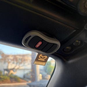 May include: A grey and black Genie garage door opener remote control fixed to the interior roof of a vehicle. The remote features three black buttons and the Genie logo in red.