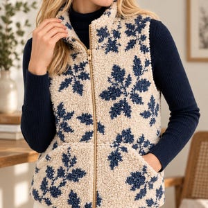 K&ouml;nnte beinhalten: Eine &auml;rmellose Rei&szlig;verschlussweste aus cremefarbenem Sherpa-Fleece mit dunkelblauem Blumenmuster. Das Kleidungsst&uuml;ck hat einen hohen Kragen, einen mittigen Rei&szlig;verschluss aus goldfarbenem Metall und zwei Vordertaschen. Der Text FamilySewingCo ist sichtbar.
