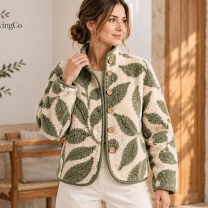 May include: A cream and sage green fleece jacket featuring a repeating leaf pattern, a high collar, and a front button closure with light brown wooden buttons. The jacket has long sleeves and a straight hemline, constructed from a textured material.