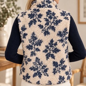 K&ouml;nnte beinhalten: Eine &auml;rmellose Weste aus cremefarbenem Sherpa-Fleece mit einem sich wiederholenden dunkelblauen Blumenmuster. Die Weste hat einen hohen Kragen und wird &uuml;ber einem dunkelblauen, lang&auml;rmeligen Rippshirt und einer wei&szlig;en Hose getragen. Der Text FamilySewingCo ist sichtbar.