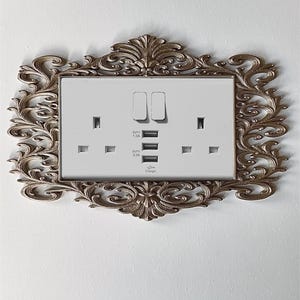 May include: A white electrical socket with two switches and USB charging ports, set within an ornate, antique-style gold frame. The frame features intricate scrollwork and decorative elements, adding a touch of elegance to the functional design.
