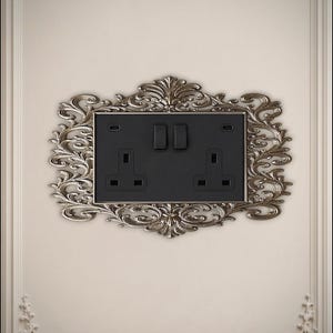 May include: A rectangular black double electrical wall socket with two switches and two USB-C ports, framed by an ornate, metallic silver-toned decorative border featuring intricate scrollwork and leaf-like patterns on a plain wall surface.