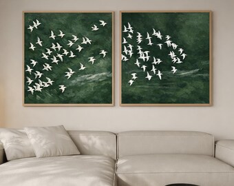Puede incluir: Dos obras de arte enmarcadas que representan pájaros blancos en vuelo sobre un fondo verde oscuro. Los marcos son de color marrón claro. La obra de arte se exhibe sobre un sofá seccional color crema y una mesa de centro de madera clara. La habitación tiene una combinación de colores neutros.