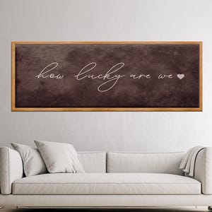 Può includere: Un'opera d'arte murale rettangolare con cornice in legno e sfondo marrone scuro. Il testo bianco in corsivo recita "how lucky are we" con un piccolo simbolo a forma di cuore. L'opera d'arte è esposta sopra un divano bianco.