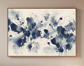 Puede incluir: Impresión artística abstracta con salpicaduras y goteos de acuarela azul marino sobre un fondo crema. La obra de arte está enmarcada en un marco fino de color dorado. La pintura se exhibe sobre un sofá beige y una mesa de madera.
