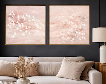 Puede incluir: Dos obras de arte enmarcadas que representan pájaros blancos en vuelo sobre un cielo rosa y melocotón. Los marcos son dorados. El arte se exhibe sobre un sofá crema con cojines beige, creando una estética relajante.