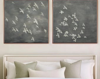 Puede incluir: Dos obras de arte murales cuadradas enmarcadas. Cada una presenta una bandada de pájaros blancos en vuelo sobre un fondo gris oscuro. Los marcos son de un color metálico cálido. La obra de arte se exhibe sobre una cama con ropa de cama blanca y cojines decorativos.