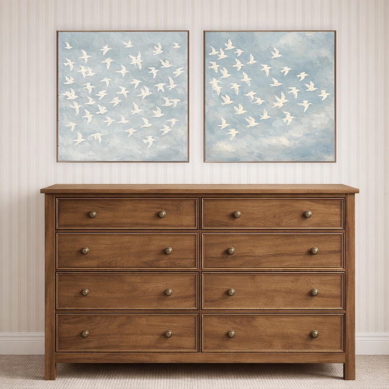 May include: Two framed artworks depict white birds in flight against a light blue sky with clouds. Below, a wooden dresser with eight drawers and brass knobs provides storage. The dresser is a warm brown color.