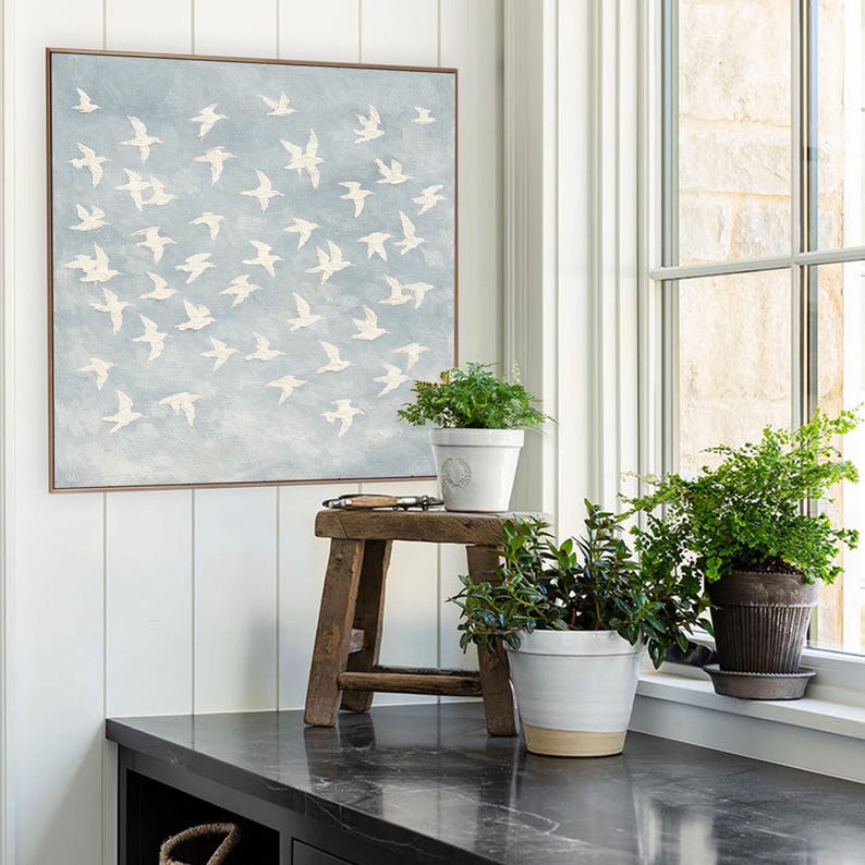 May include: Square framed artwork featuring a flock of white birds against a light blue sky. The artwork is displayed above a dark countertop with potted plants and a small wooden stool. The scene is set near a window.