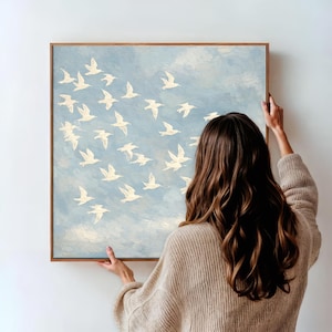 May include: A square painting with a light blue background and a flock of white birds in flight. The painting is in a wooden frame. The painting is being held by a person.