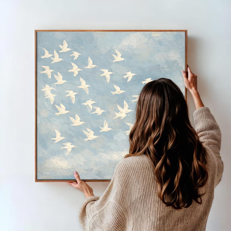 May include: A square painting with a light blue background and a flock of white birds in flight. The painting is in a wooden frame. The painting is being held by a person.