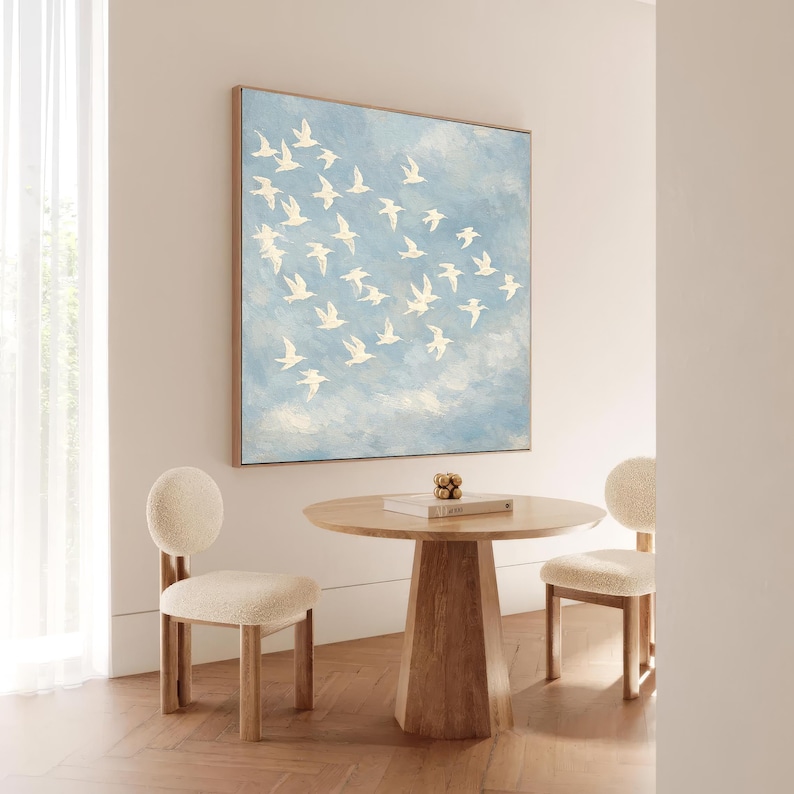 May include: A framed artwork depicts white birds in flight against a light blue sky. A round wooden table and two chairs with rounded backs and cream-colored upholstery are in front of the artwork. A decorative object and a book are on the table.
