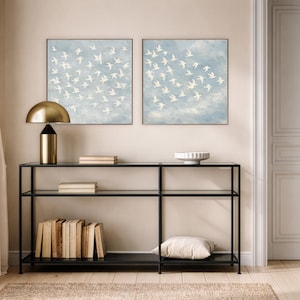 May include: Two framed artworks depict white birds in flight against a light blue sky. A black metal console table with glass shelves holds books, a gold lamp, and a white bowl. A beige rug and a white pillow complete the scene.