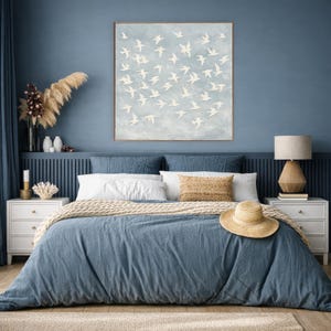 May include: A bedroom scene with a blue comforter and pillows. A large framed artwork of white birds against a light blue background hangs above the bed. A straw hat rests on the comforter, and a wooden lamp sits on a nightstand.