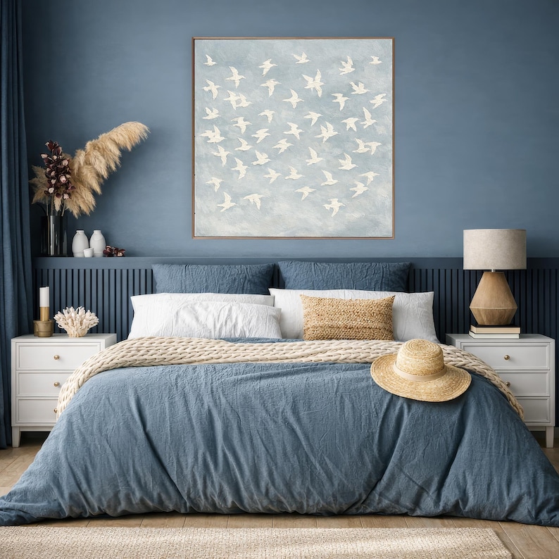 May include: A bedroom scene with a blue comforter and pillows. A large framed artwork of white birds against a light blue background hangs above the bed. A straw hat rests on the comforter, and a wooden lamp sits on a nightstand.