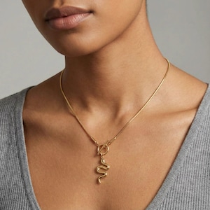 Può includere: Una collana color oro con un ciondolo a forma di serpente. La collana ha una catena sottile e una chiusura a levetta. Il ciondolo a forma di serpente è dettagliato con un design curvo. La collana è indossata su un top grigio.