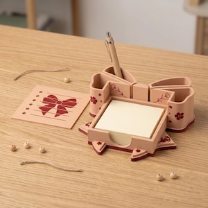 May include: A pink desk organizer shaped like a bow, with compartments for pens and sticky notes. A wooden pen sits in one compartment. A matching notepad with a bow design is also present.