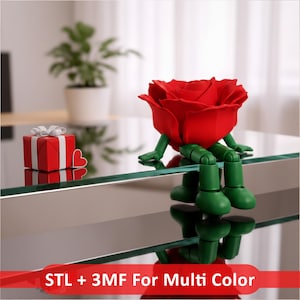 Puede incluir: Una figura de rosa roja con brazos y piernas verdes articulados sentada en un borde, acompañada de un pequeño regalo rojo con cinta blanca y un corazón rojo. El texto en la parte inferior dice STL + 3MF For Multi Color.