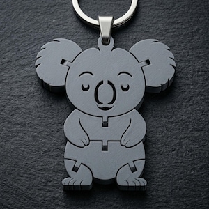 Può includere: Un portachiavi grigio a forma di koala con un portachiavi argentato. Il koala ha una faccia sorridente con gli occhi chiusi e un design dettagliato a strati. Il portachiavi è realizzato in un materiale solido.