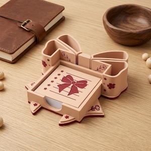 May include: A light brown desk organizer shaped like a bow, with compartments for pens and a notepad. The notepad has a red bow design. The organizer is decorated with red floral accents and sits on a wooden surface.