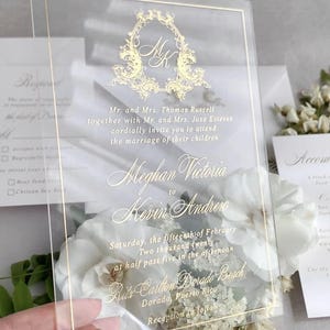 Könnte beinhalten: Klare Hochzeitskarte aus Acryl mit goldfarbenem Schriftzug und goldenem Rand. Die Einladung enthält die Namen Meghan Victoria und Kevin Andrew sowie das Hochzeitsdatum und den Ort. Die Einladung ist von weißen Blumen umgeben.