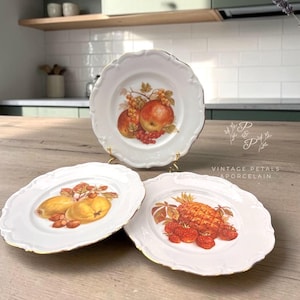 May include: Three white porcelain plates with scalloped edges and gold trim. The top plate features a red apple and grapes. The bottom left plate shows yellow pears and nuts. The bottom right plate displays a pineapple and red strawberries.