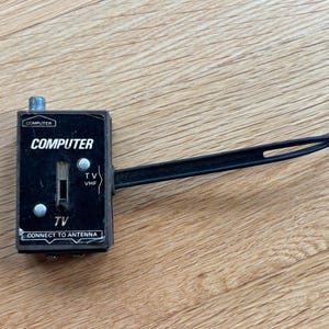 May include: A black rectangular electronic switch box with a sliding toggle, featuring white text that reads COMPUTER, TV, and CONNECT TO ANTENNA. It includes a long black cable with a two-pronged metal spade connector at the end for signal input.