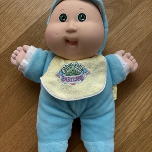 May include: A Cabbage Patch Kids doll with a round face, green eyes, and a soft vinyl head. The doll wears a light blue plush onesie with a hood and a yellow bib featuring the embroidered text Cabbage Patch Kids BabyLand.